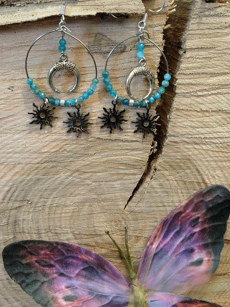 boucles-d-oreilles-atlante-en-apatite-bleue-creation-magic-gaia-suspendues-sur-bois-clair-details-lune-et-soleils-decor-papillon-en-bas-apaisement-clarte-expression.jpeg Boucles d’oreilles atlante apatite - création magic gaïa – Image 1