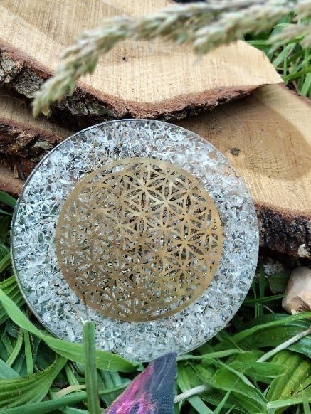 plateau-de-rechargement-en-orgonite-transparent-a-eclats-blancs-motif-fleur-de-vie-dore-au-centre-pose-dans-l-herbe-avec-rondins-de-bois-en-decor-favorise-purification-apaisement-et-harmonie.jpeg Plateau orgonite cristal de roche fleur de vie laiton – Image 1