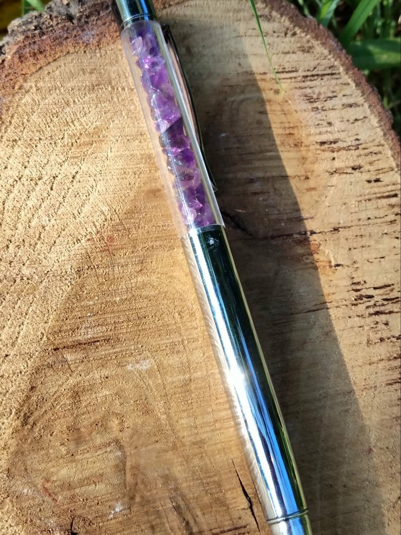 stylo-amethyste-rechargeable-pose-sur-une-tranche-de-bois-en-exterieur-tube-transparent-rempli-d-eclats-violets-ecriture-fluide-et-design-elegant.jpeg Stylo améthyste – Image 1